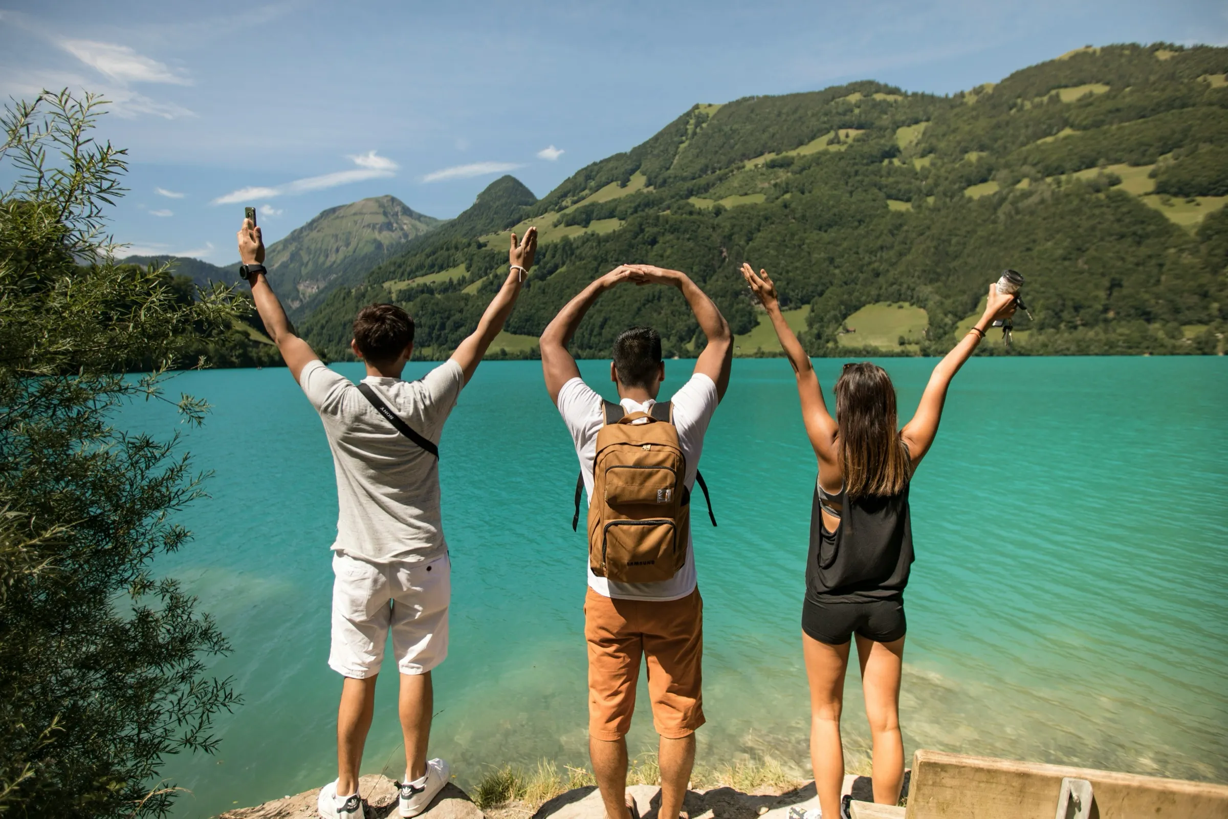 Three travelers cheering with their arms raised beside a turquoise lake