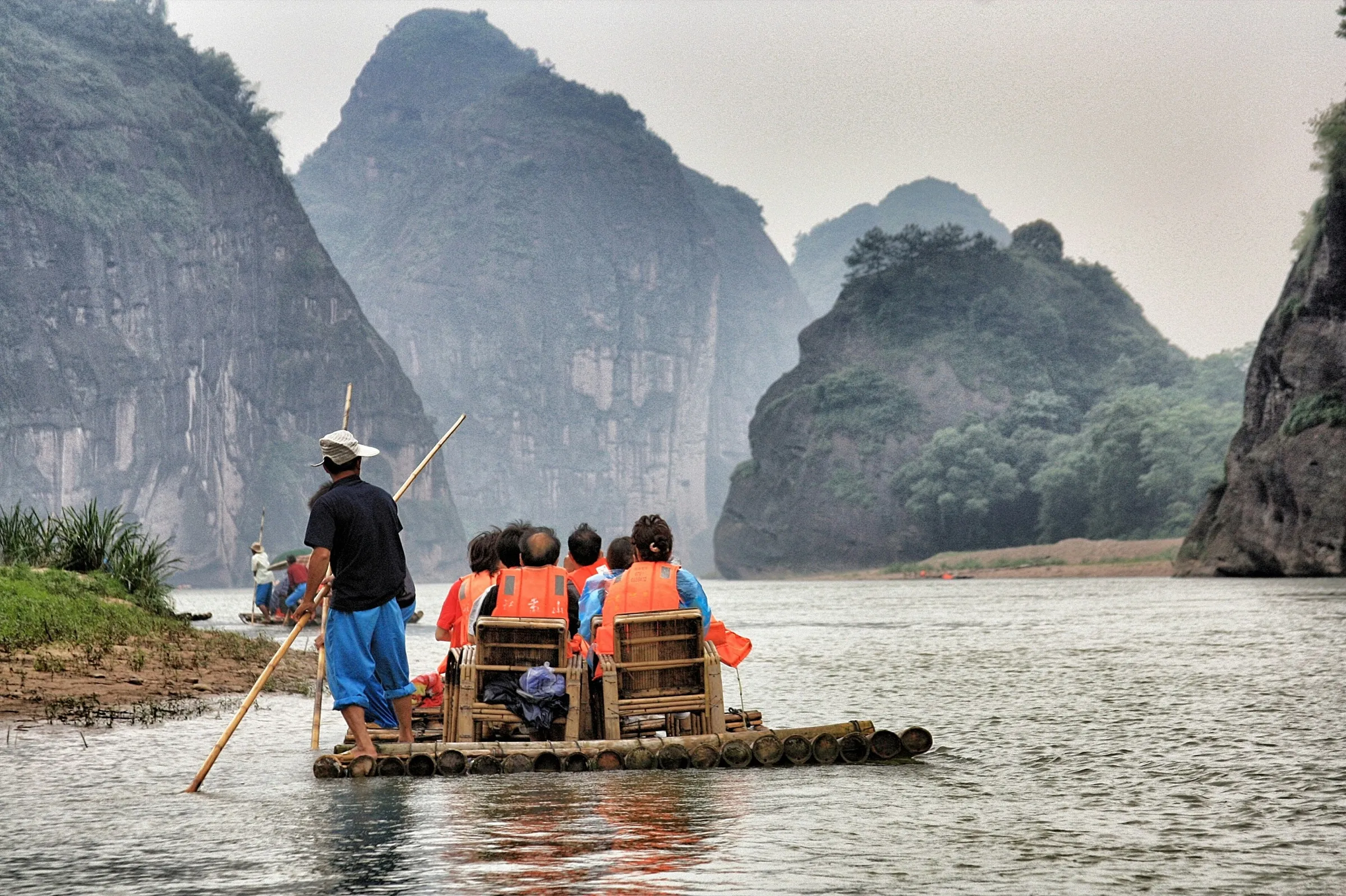 A guide poling a bamboo raft of travelers down a river between karst mountains